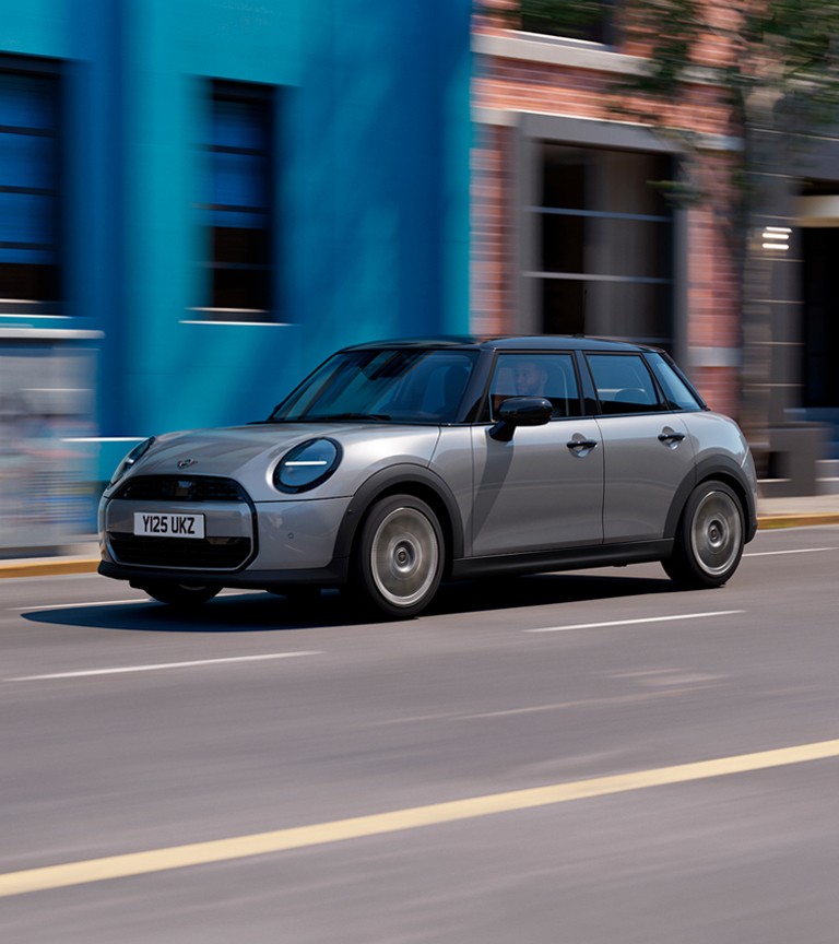A silver MINI Cooper 5-Door drives swiftly down a city street, passing a bright blue wall and brick building. The scene conveys a sense of speed and urban energy.